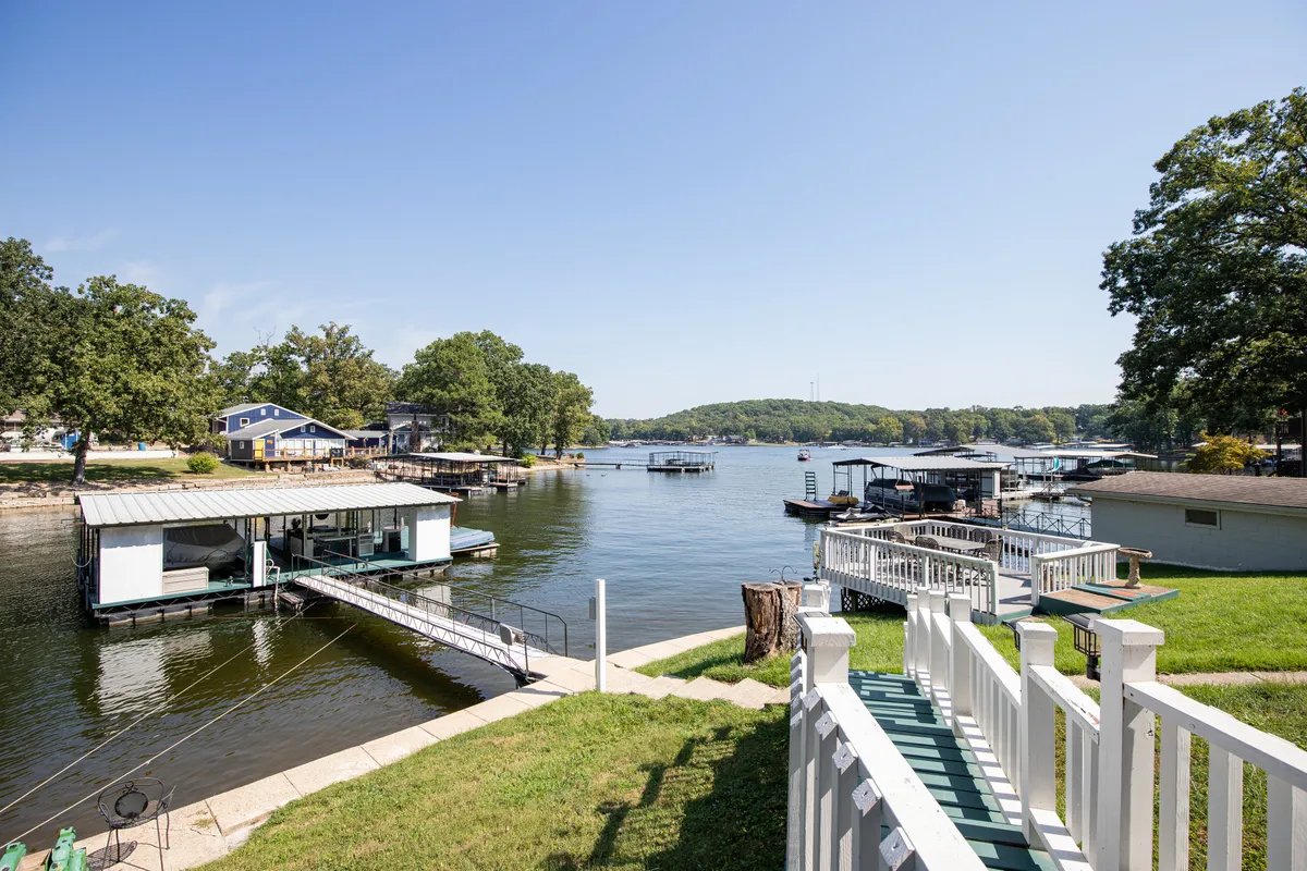 A wide view showcasing the serene lake, boathouses, and a waterfront dock surrounded by greenery. The calm water, visible dock, and accessible shoreline evoke a classic fishing scene and highlight the connection between the home and the lake itself.