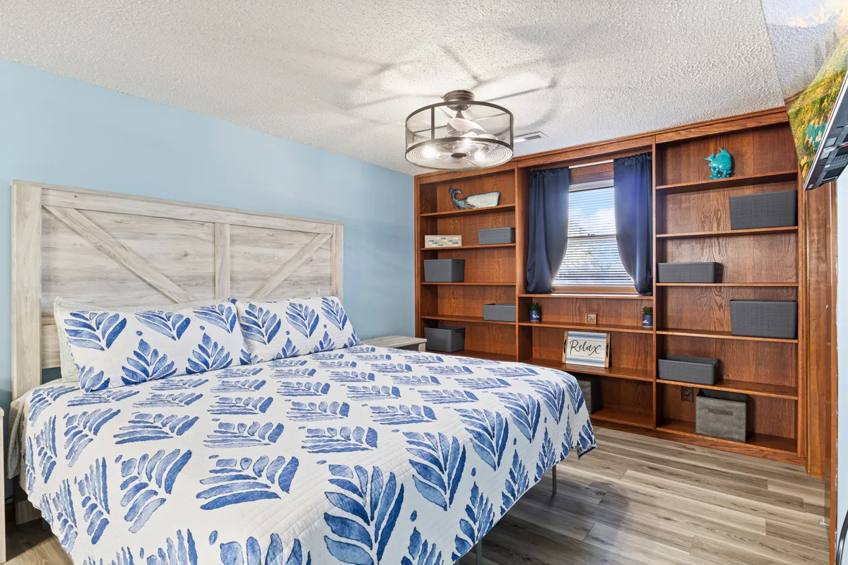 A cozy bedroom showcasing a bed with a blue leaf-patterned quilt, a wooden shelving unit, and decorative items artfully arranged on the shelves. The room is calm and inviting, perfect for unwinding after a busy day.