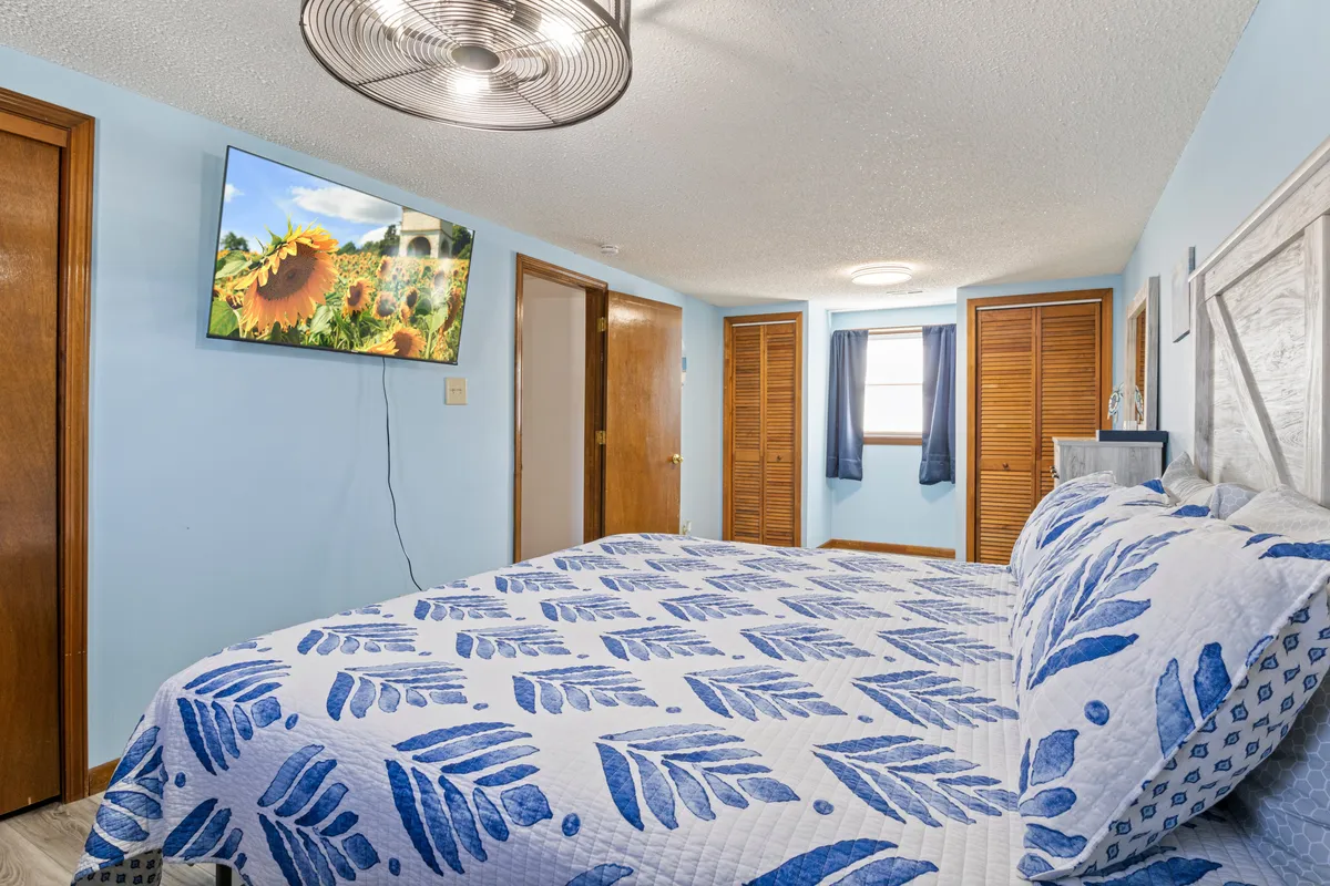 A welcoming bedroom with a bed dressed in a blue and white leaf-patterned quilt, a television mounted on the wall, and wooden doors. Natural light streams in, creating a bright, inviting space for both rest and leisure.