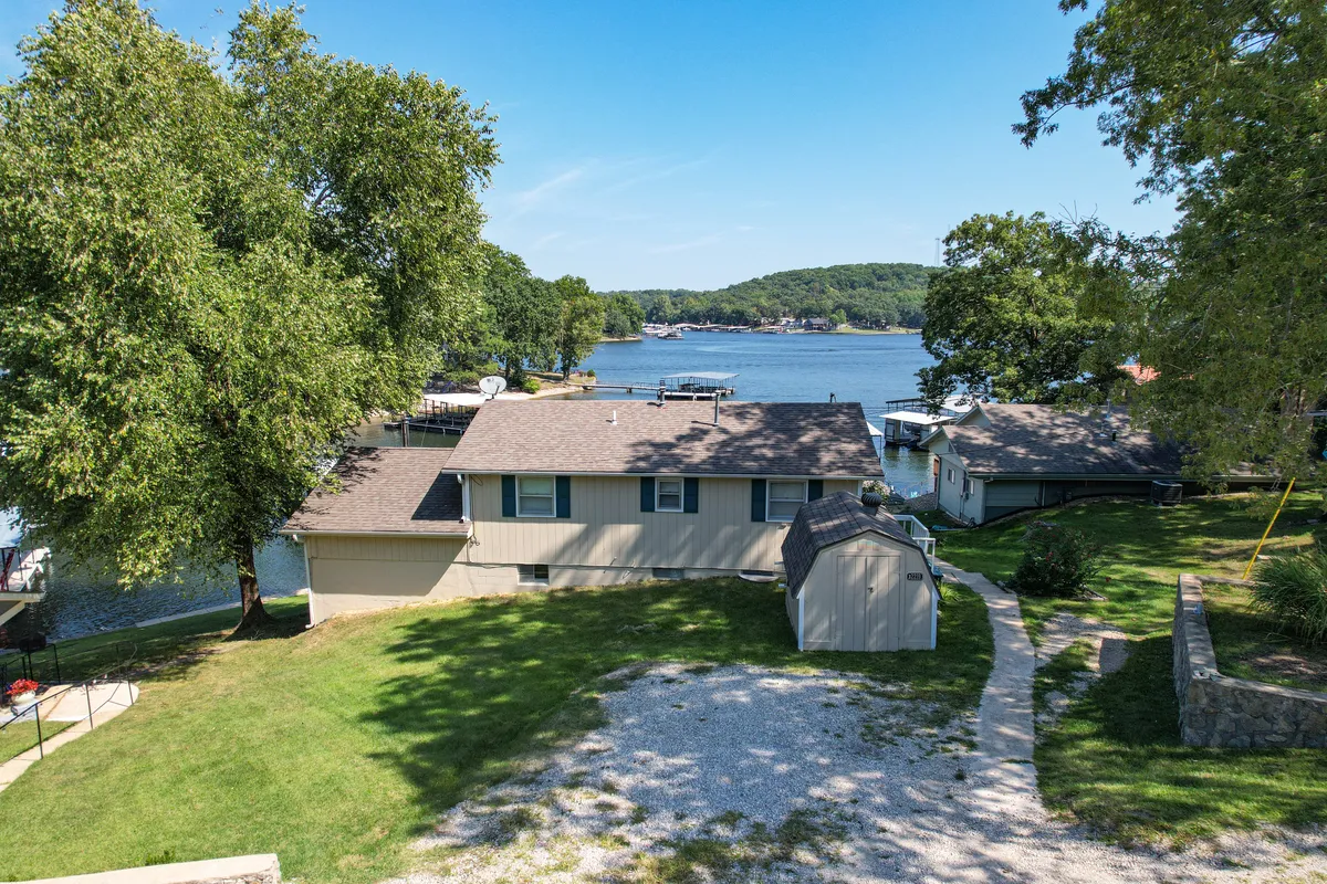 This exterior image captures the house nestled among trees, its private dock extending over the tranquil waters of Lake of the Ozarks. The scene suggests both accessibility to the water and a peaceful setting for fishing, particularly with the dock clearly visible and inviting for early morning or afternoon relaxation.