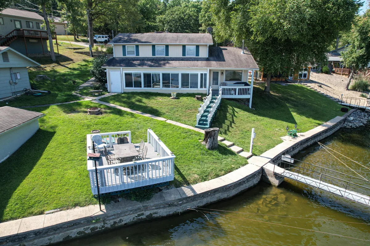 A wide view of the home's lakefront deck area with steps leading down to the water, set against a backdrop of large windows and shaded by mature trees. The inviting outdoor space is well-suited for family gatherings, dining, and easy access to boating, fishing, or swimming in the gentle cove.