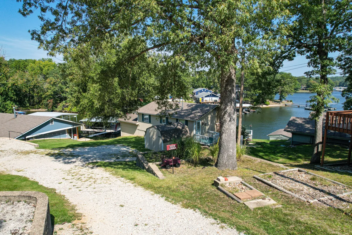 A picturesque exterior shot with a large tree shading a path leading to the house, the lake in the background, and neighboring homes visible nearby. The scene feels welcoming and hints at both relaxation and local exploration options.