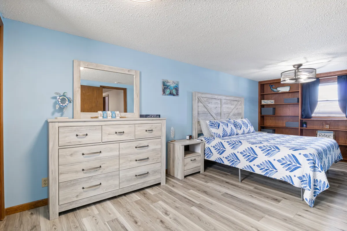 A tranquil bedroom featuring a bed with blue leaf-patterned bedding, a light wood dresser, a decorative mirror, and an open shelving unit. The space is lit with soft natural light, and subtle coastal accents add to the serene vibe.