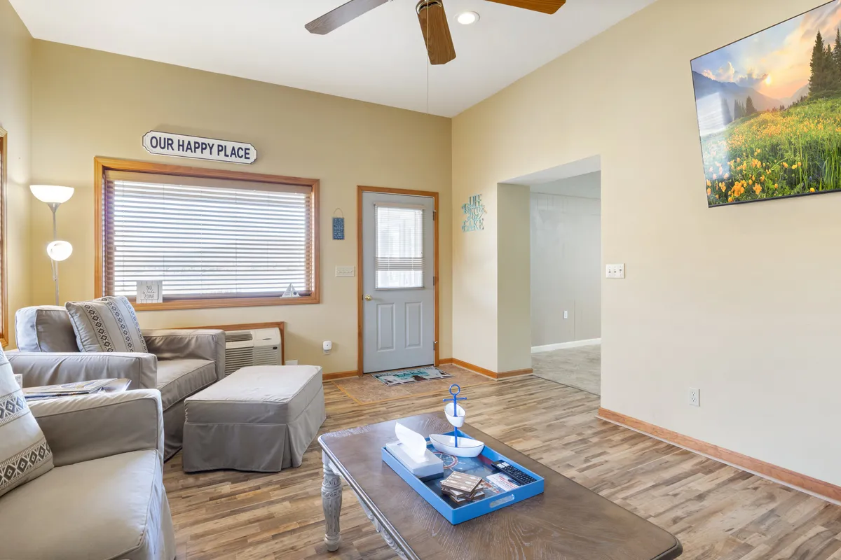 A bright, cheerful living room interior with a sofa, coffee table, TV, and large windows letting in natural light. The space is designed for relaxing, socializing, watching TV, and group activities like game nights.