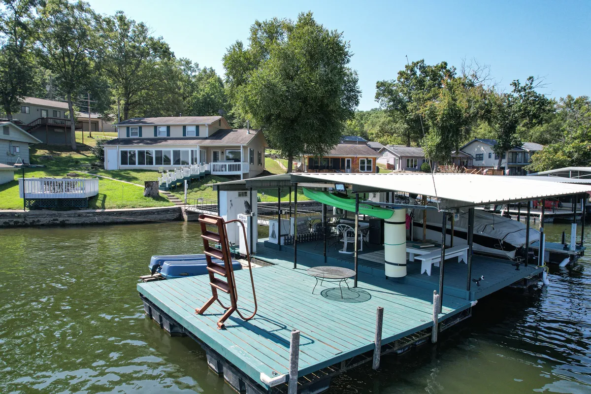The image showcases a serene lakefront setting with a spacious dock extending over calm cove waters, surrounded by lush green trees and shoreline homes. Kayaks are visible on the dock, inviting guests to explore the water, while the gentle lot and comfortable seating areas make it ideal for relaxing, fishing, or enjoying the sunset with friends and family.