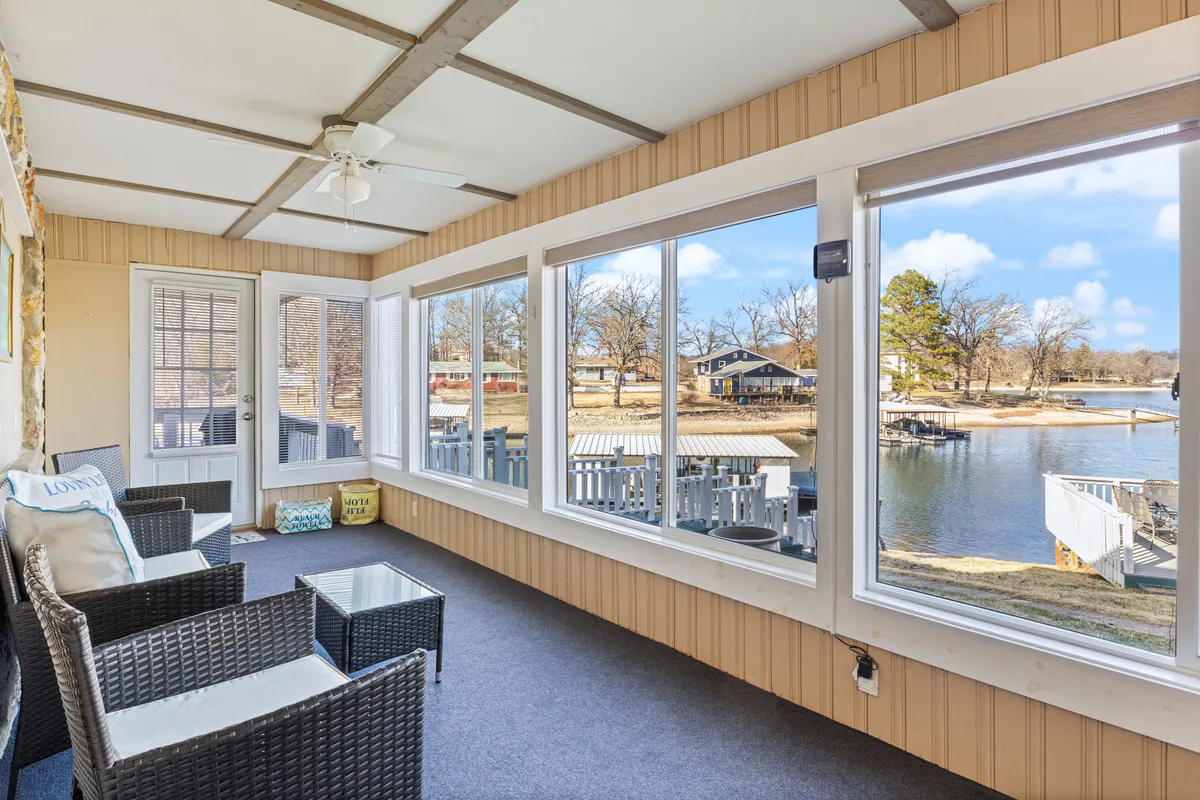 A sunroom or enclosed sitting area with large windows overlooking the water, featuring comfortable seating and decorative cushions. The view captures the serene lake, inviting relaxation and moments of reflection after outdoor fun.