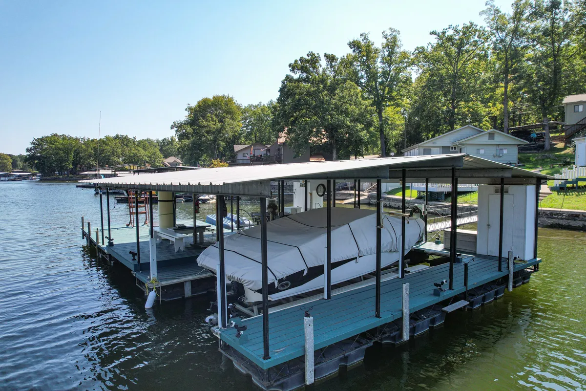 A wide view of the dock and surrounding lake, with a boat, lush greenery, and homes in the distance. The scene captures a sense of fun and recreational activity, with ample space for family gatherings and water sports.