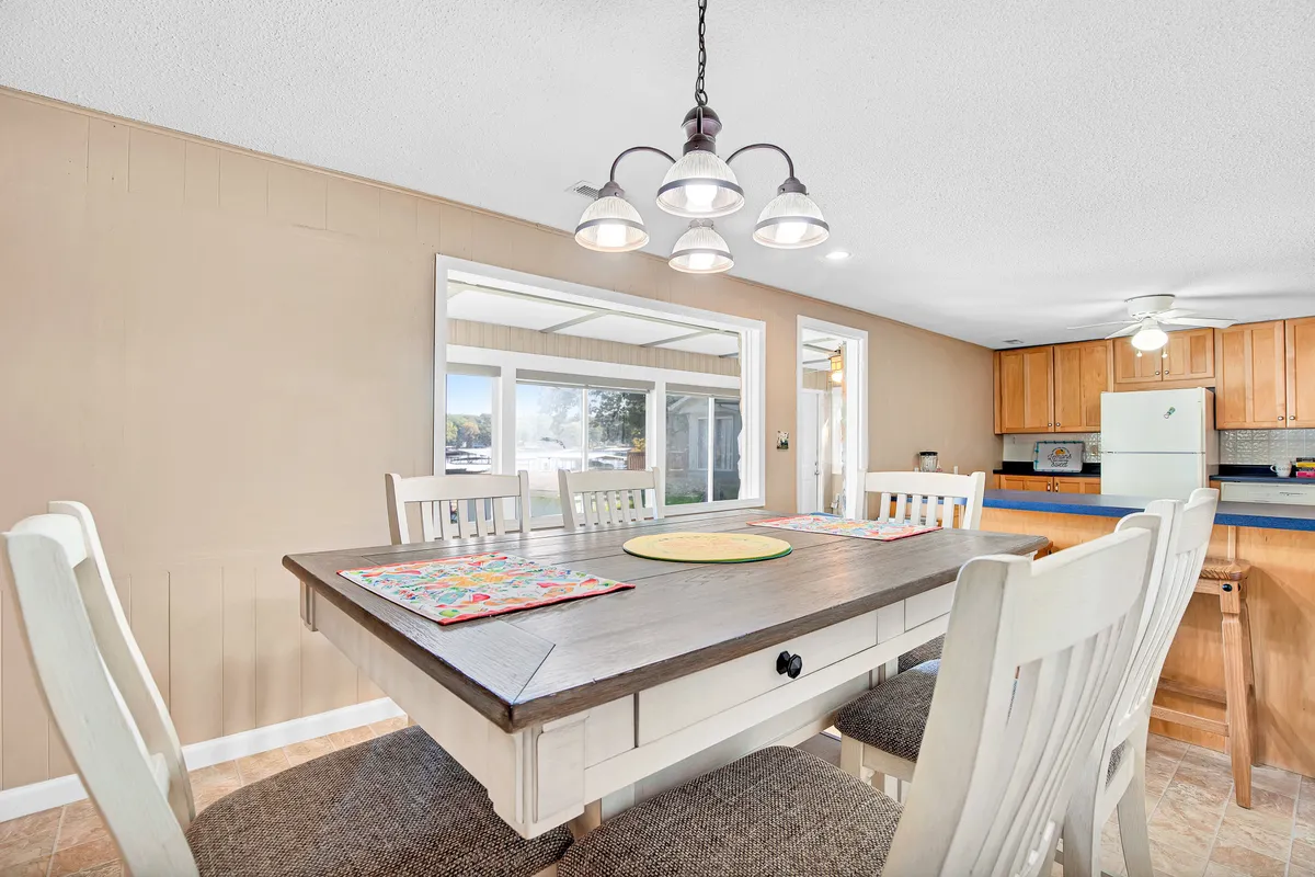 A welcoming dining area with a family-sized table, warm chandeliers, a kitchen area, and windows that fill the space with natural light. The setup is ideal for shared meals, game nights, or gathering on rainy days.